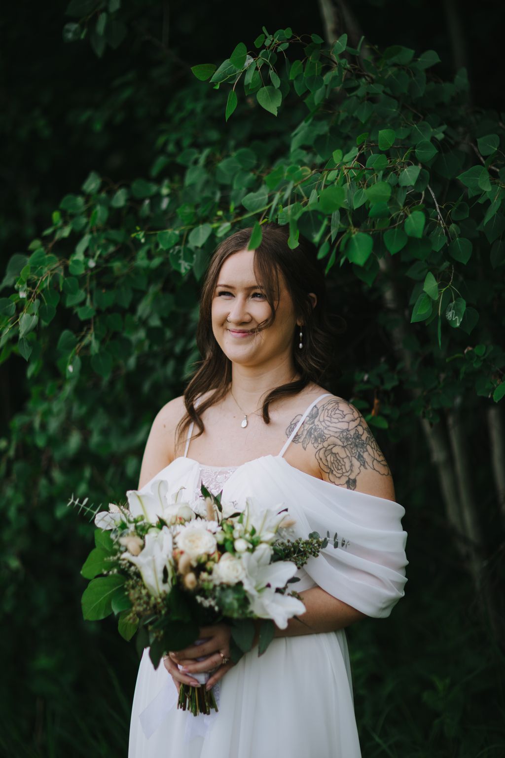 Bride in white dress holding white floral bouquet smiles beneath hanging green foliage in natural light