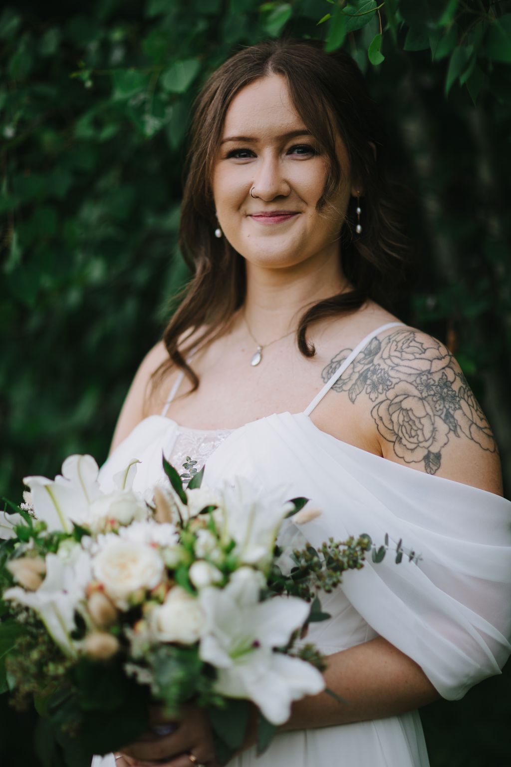 Bride in white off-shoulder dress holding white bouquet smiles outdoors against green foliage backdrop