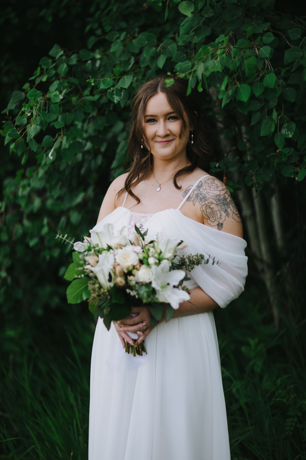 Bride in white off-shoulder dress holding white floral bouquet against lush green foliage backdrop, smiling at camera