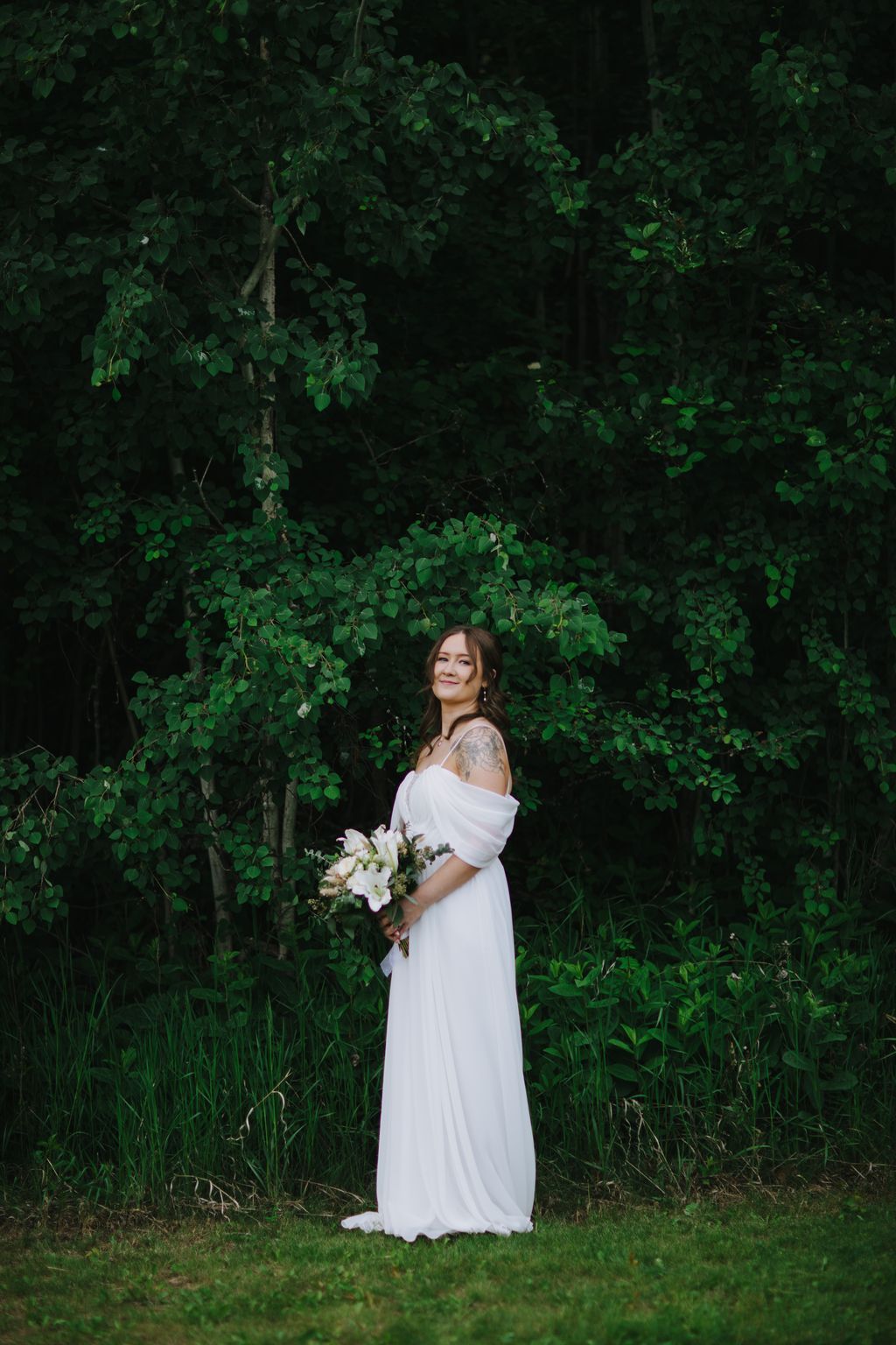 Bride in flowing white off-shoulder gown holds white bouquet against lush green ivy backdrop and lawn