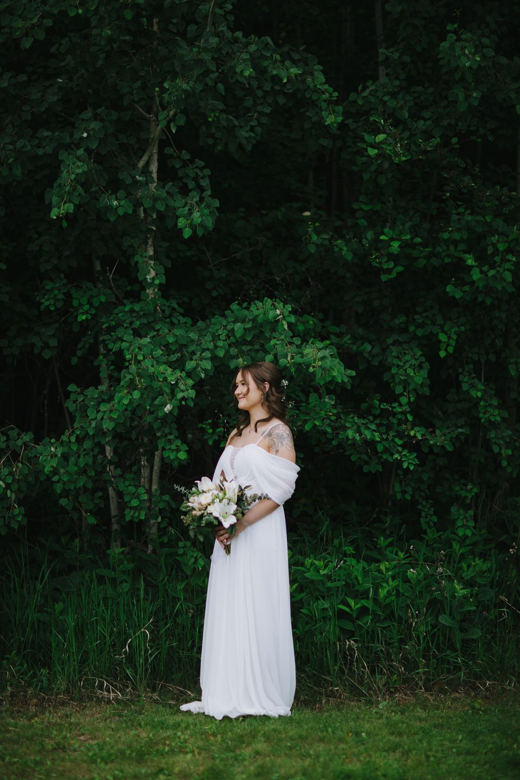 Bride in flowing white off-shoulder gown holding white flower bouquet against lush green foliage backdrop
