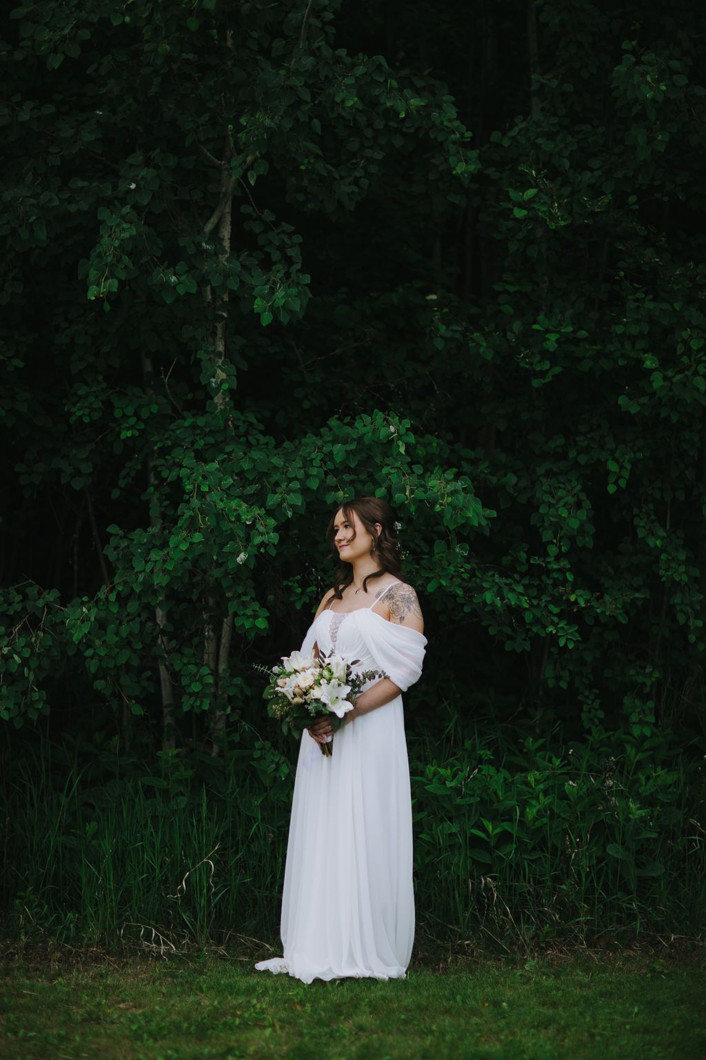 Bride in flowing white off-shoulder gown holding white bouquet stands before lush green foliage backdrop