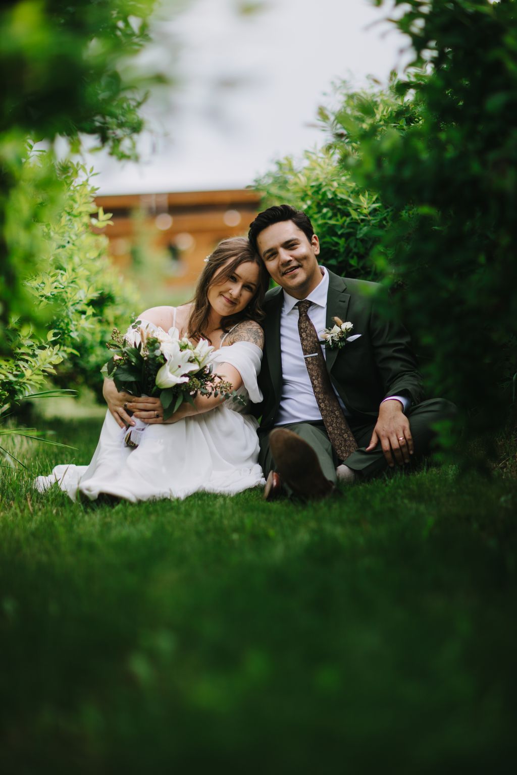 Bride in white dress holding white bouquet and groom in dark suit with patterned tie sit close together on grass surrounded by green foliage in soft natural…