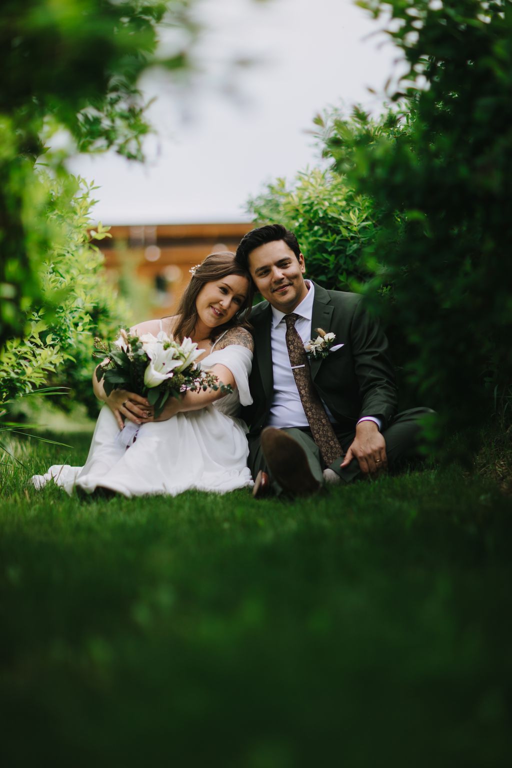 Bride and groom sitting on grass surrounded by lush green foliage, bride in white dress holding white floral bouquet, groom in dark suit with patterned tie,…