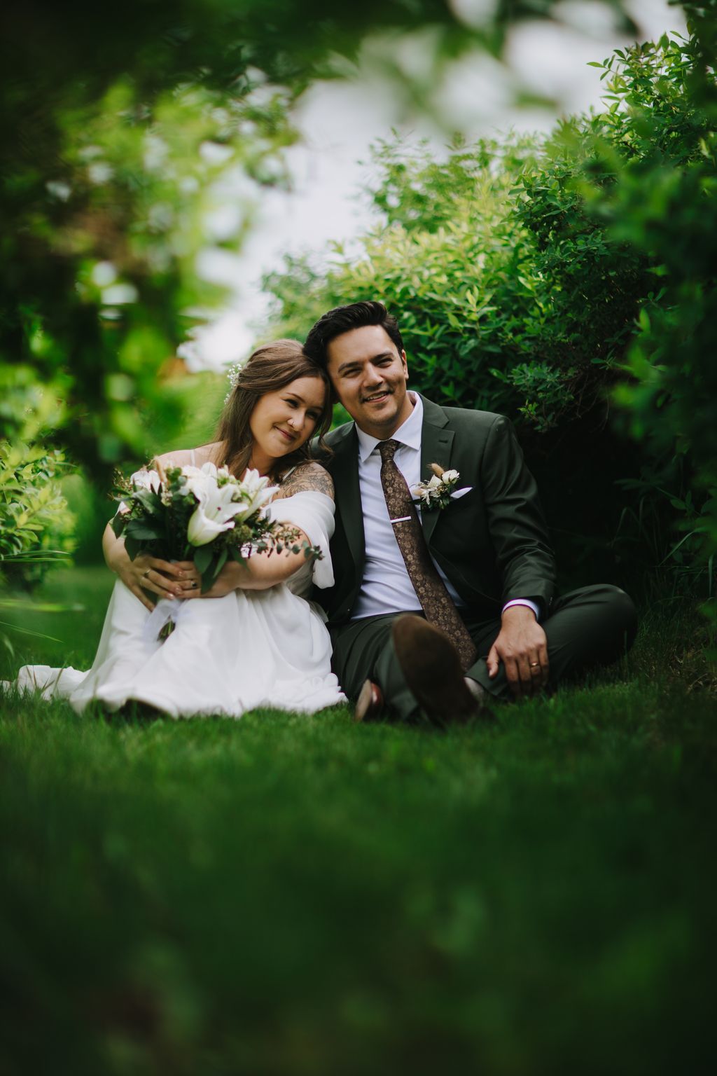 Bride in white dress and groom in olive suit sit smiling on grass surrounded by lush green foliage and white flowers