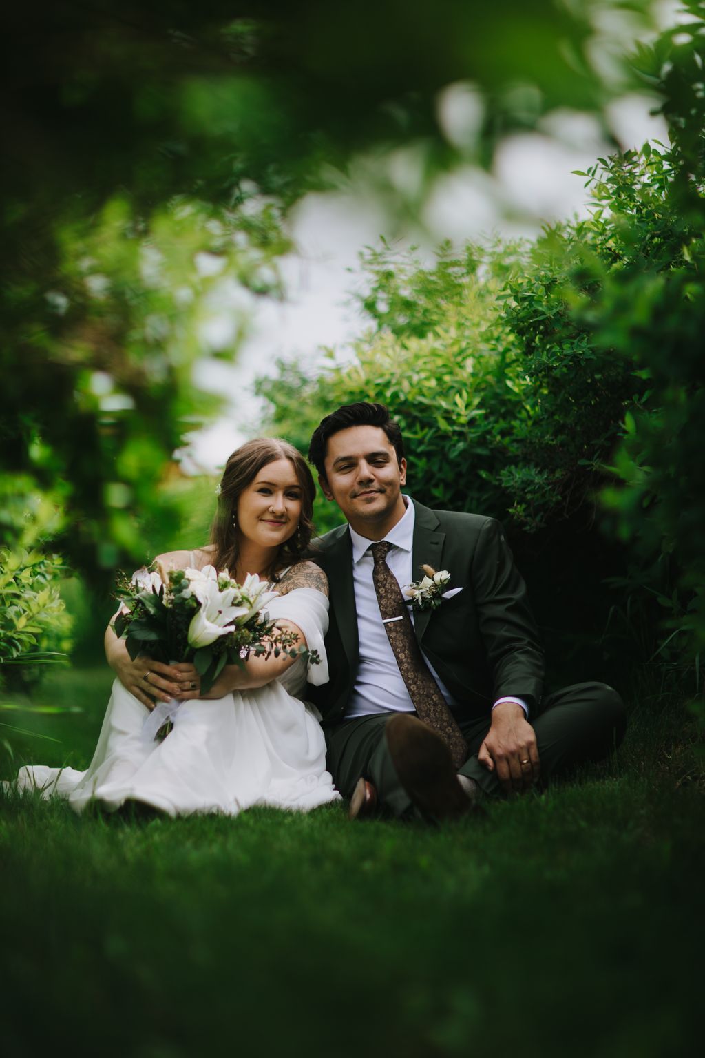 Bride in white dress holding white flowers and groom in dark suit sit together on grass surrounded by lush green foliage and blooming white flowers