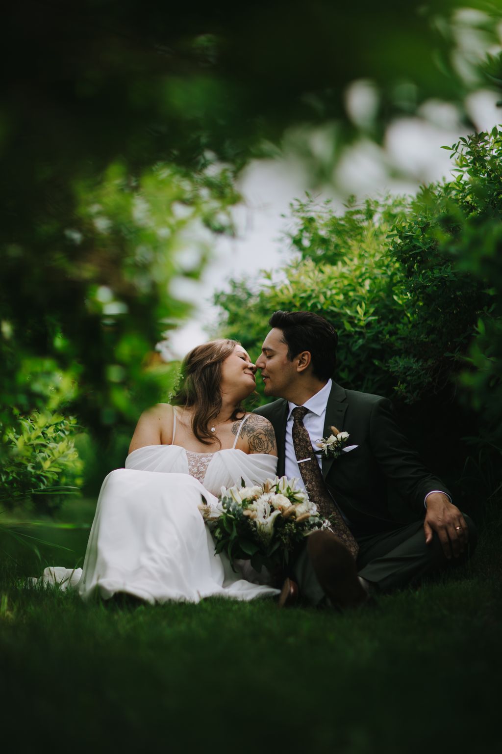 Bride and groom embrace intimately in a lush garden, holding a white bouquet, with soft green foliage and white flowers blurred in the background
