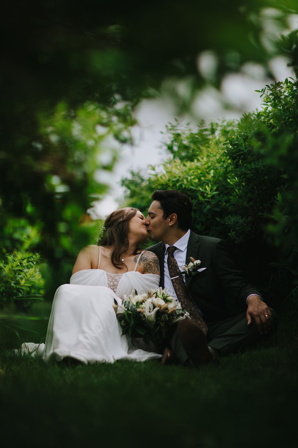 Bride and groom kiss in a lush garden with white flowers and greenery, soft natural light filtering through foliage