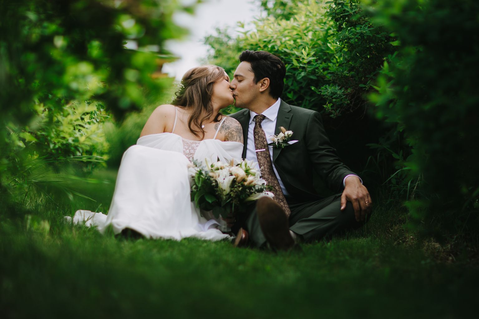 Bride and groom kiss outdoors on grass with white bouquet and lush green foliage backdrop