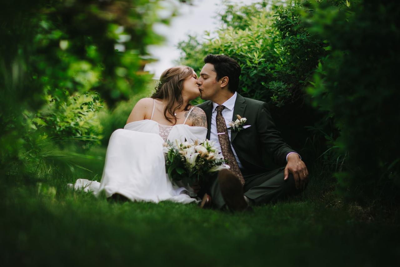 Bride and groom kiss on manicured grass with white bouquet and green hedges framing them at their Prince George wedding