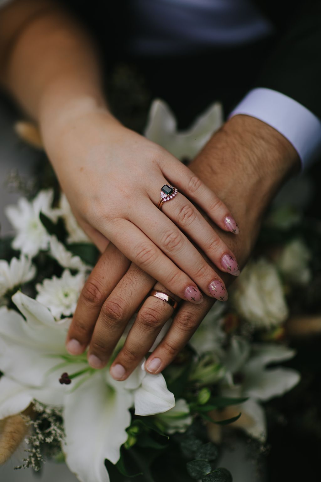 Bride and groom''s hands displaying engagement and wedding rings over white flowers in soft natural light