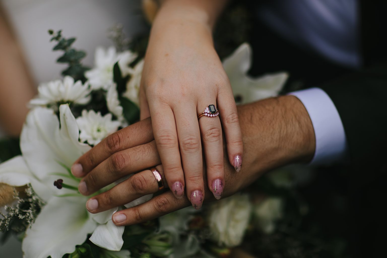 Couple''s hands together displaying matching wedding bands and an engagement ring with black stone and pink accents, with manicured nails against soft outdoor…