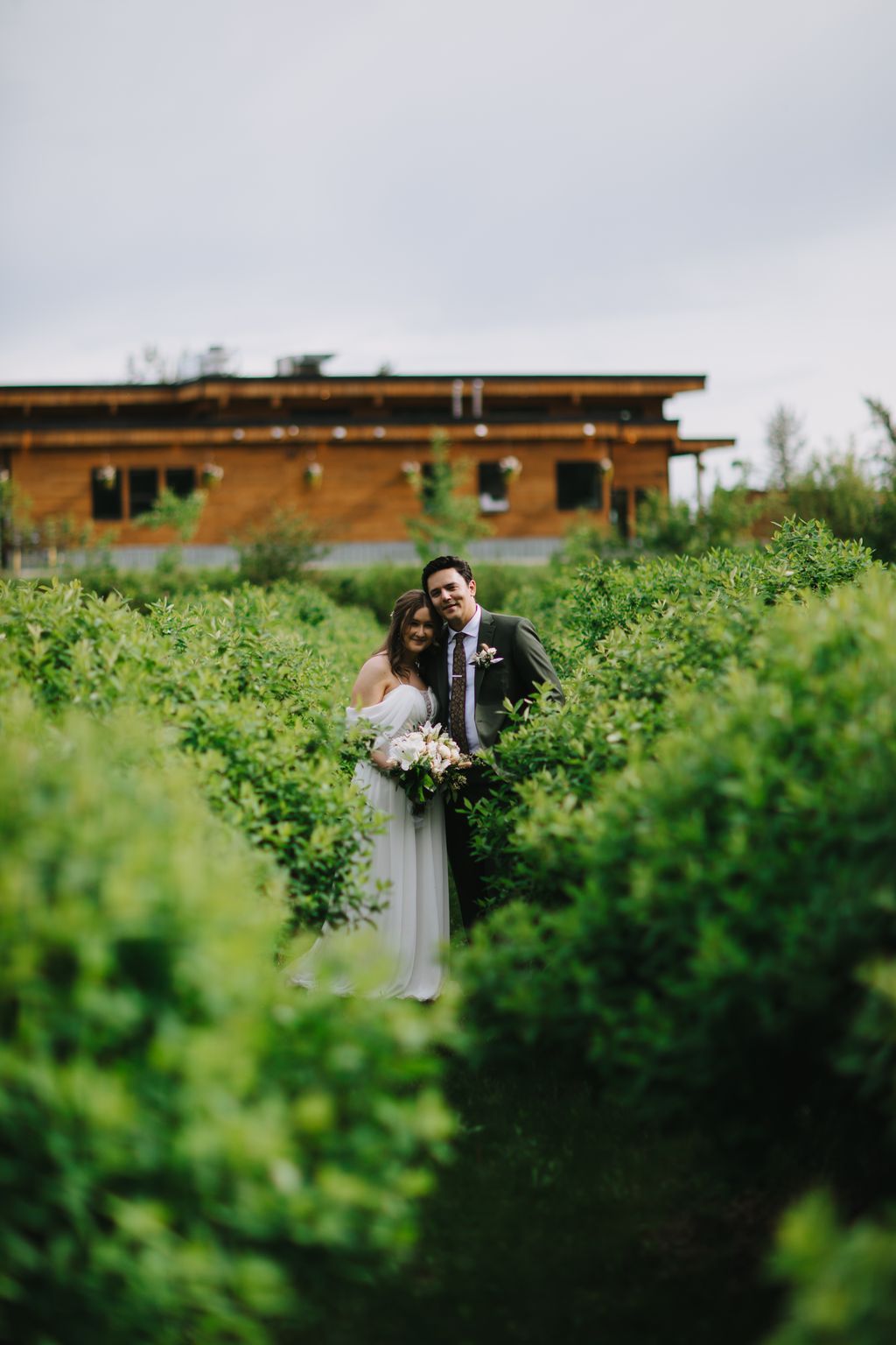 Bride and groom stand together in a lush green garden with a rustic wooden building in the background under overcast skies