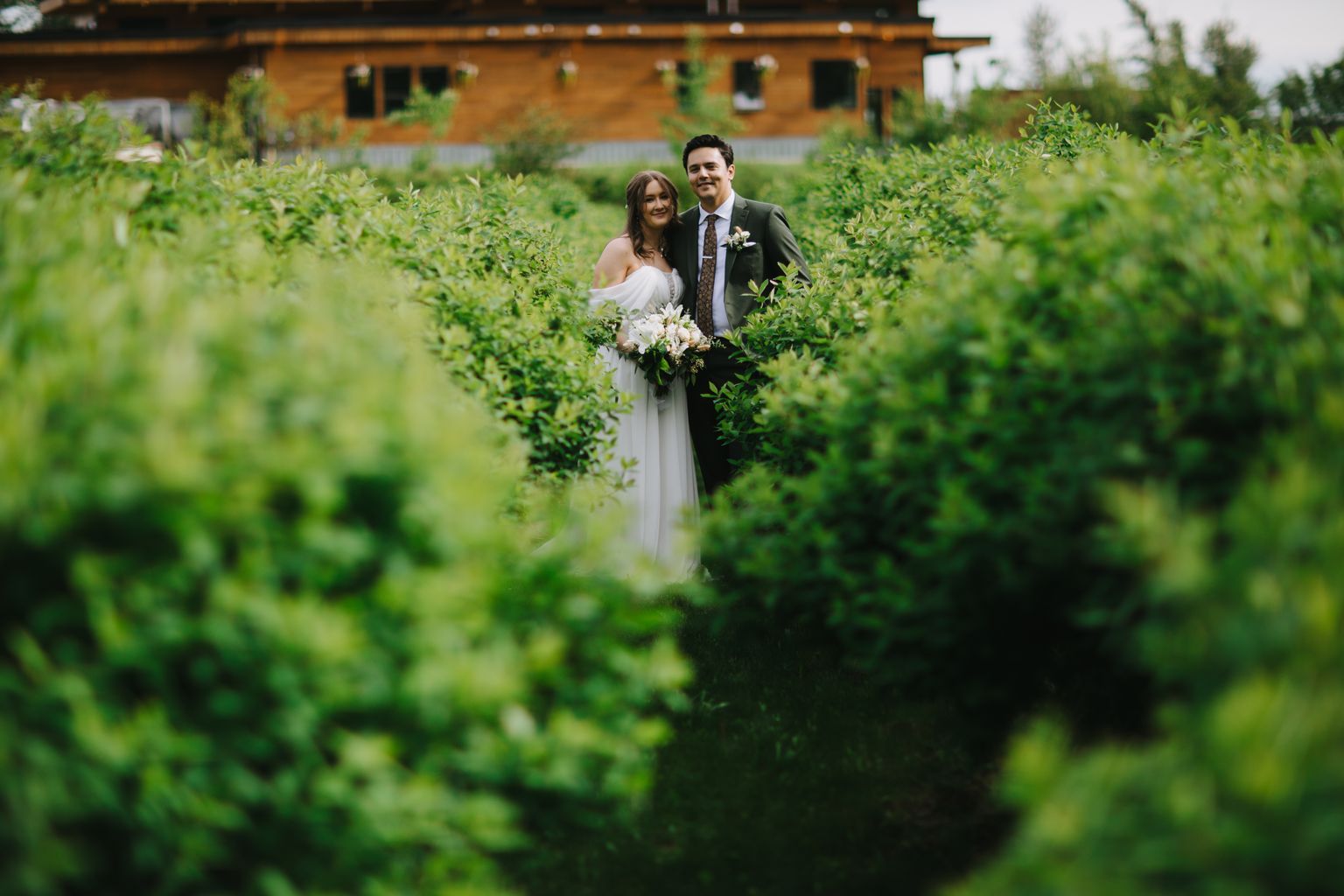 Bride in white off-shoulder gown and groom in olive suit stand together in lush green hedges, holding white bouquet, brick building behind them