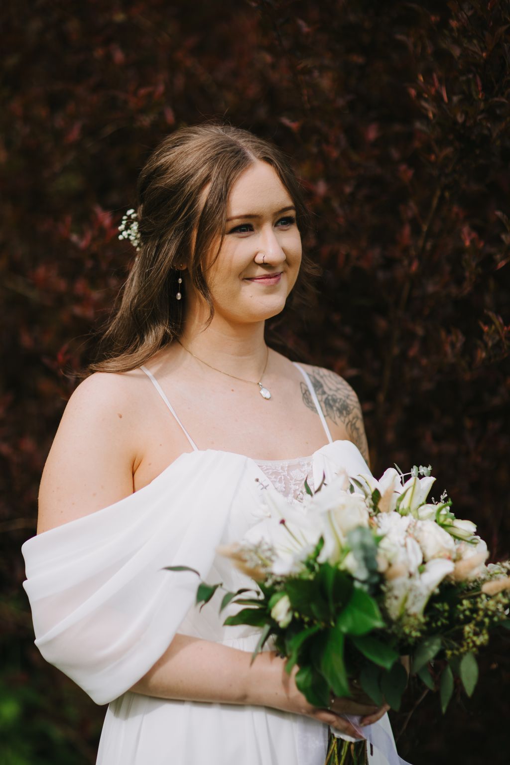 Bride in white lace dress holding white bouquet, smiling softly against dark foliage backdrop