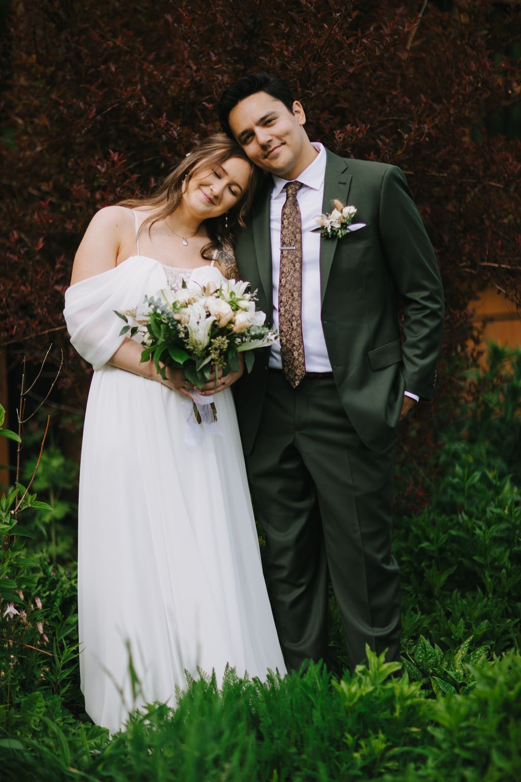 Bride and groom embrace against burgundy foliage, bride holding white bouquet, warm natural light