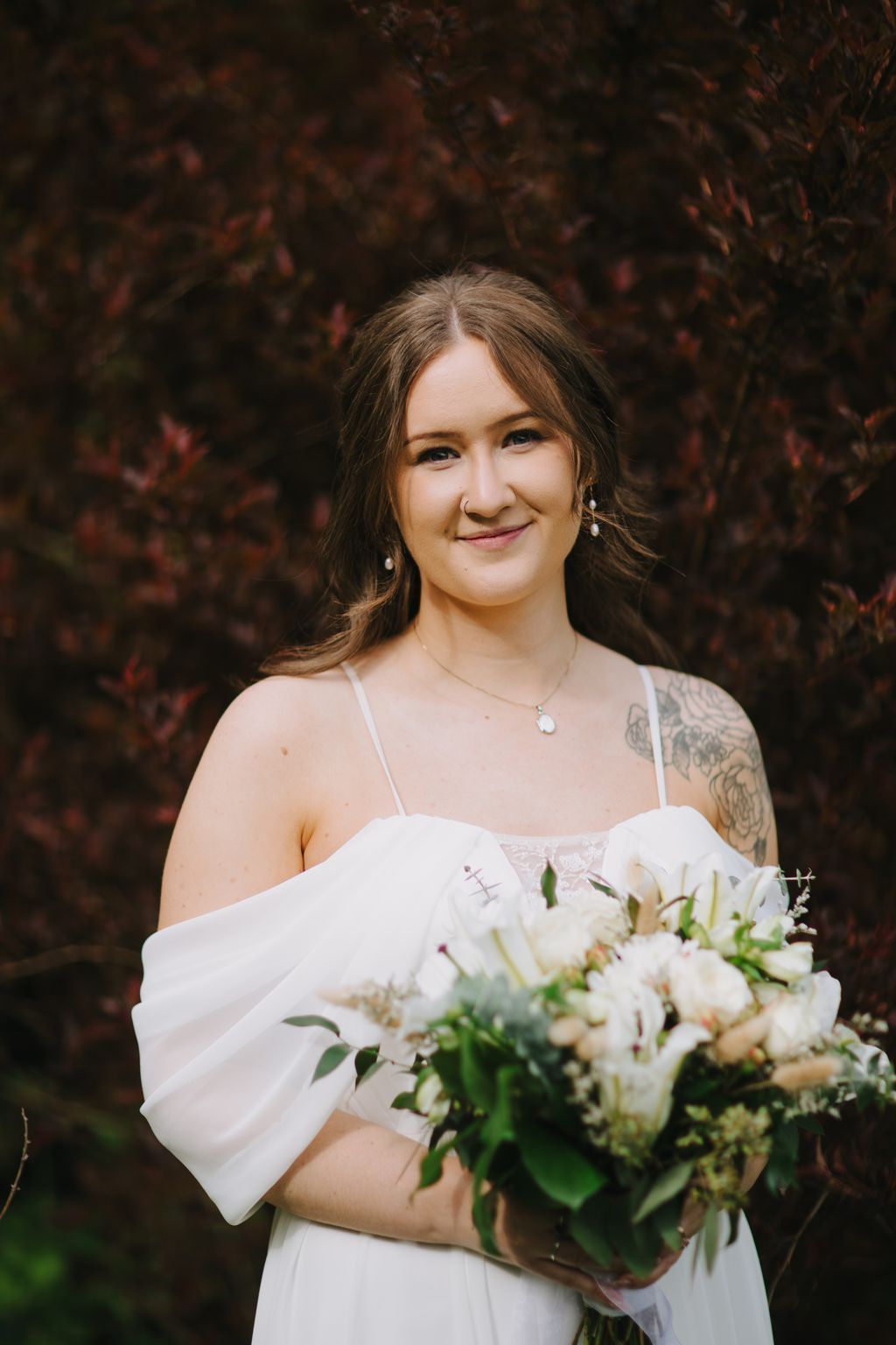Bride in white dress holding white bouquet smiles at camera against dark foliage backdrop