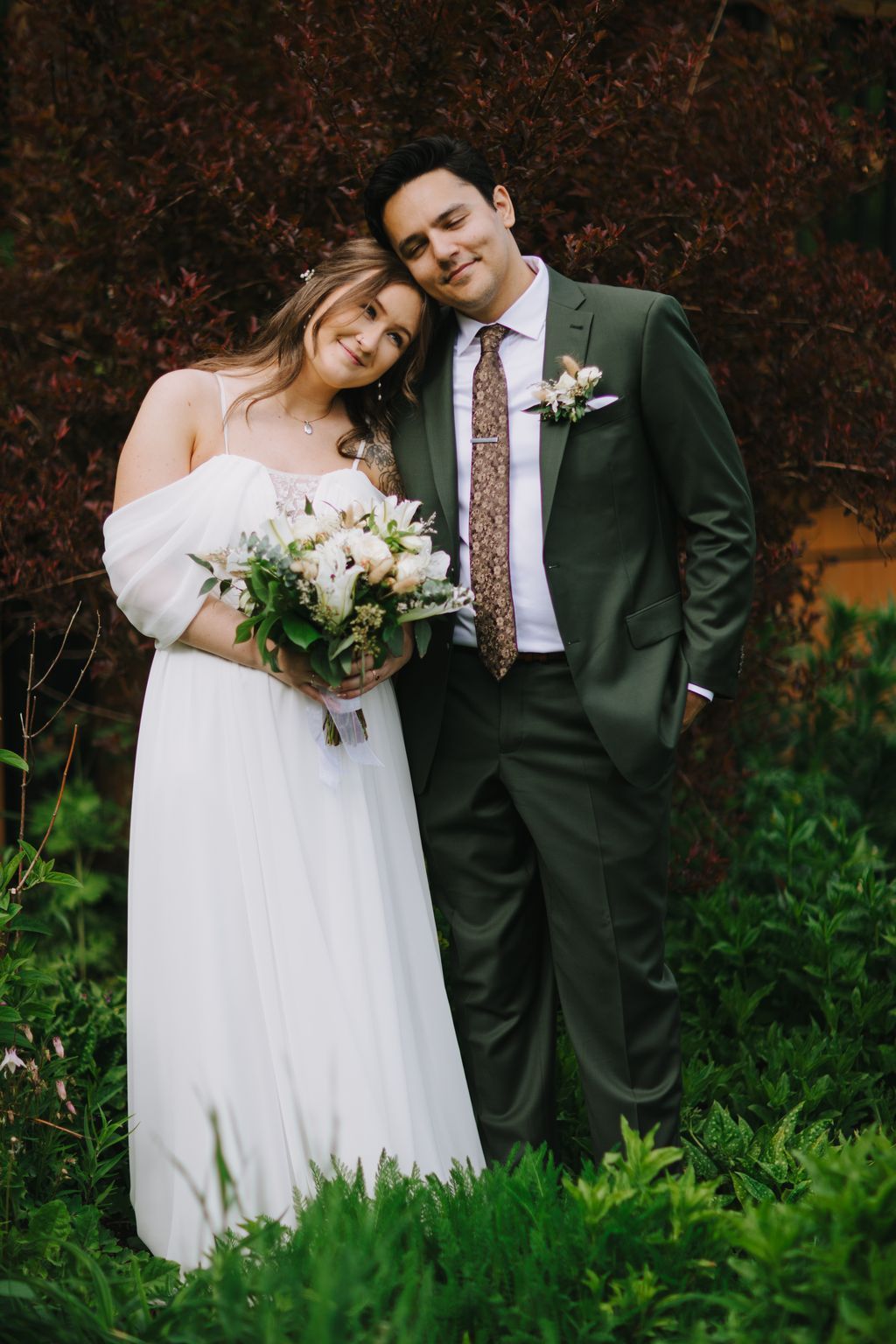 Bride in white dress and groom in sage green suit embrace against burgundy foliage, holding white bouquet, soft natural light