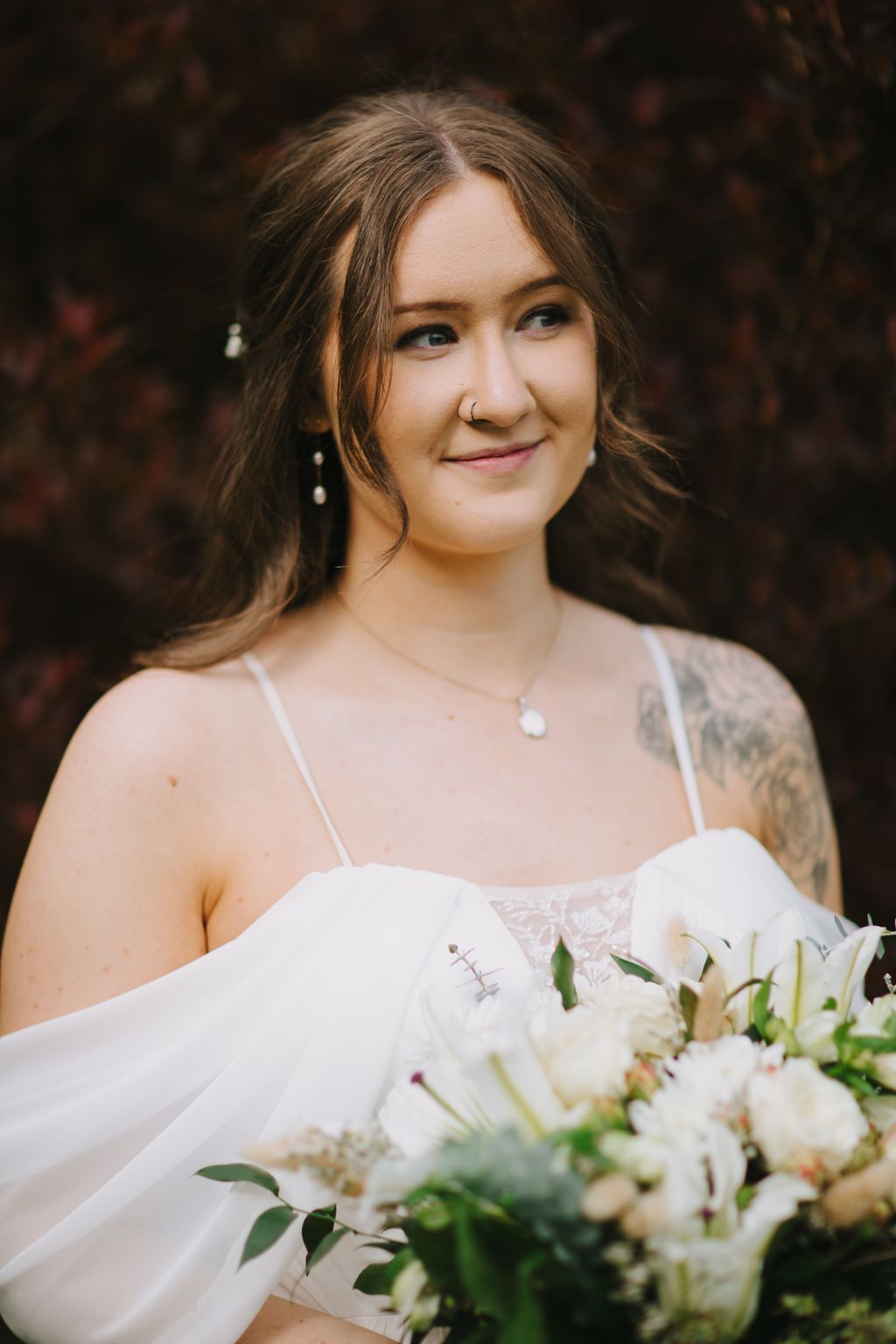 Bride in white lace dress holding white flowers, smiling softly with natural outdoor lighting and blurred background