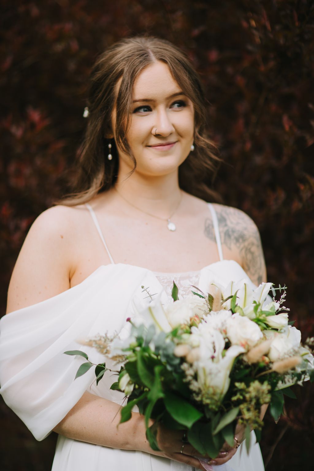 Bride in white dress holding white bouquet with greenery, smiling softly while looking away, outdoor setting with dark blurred background