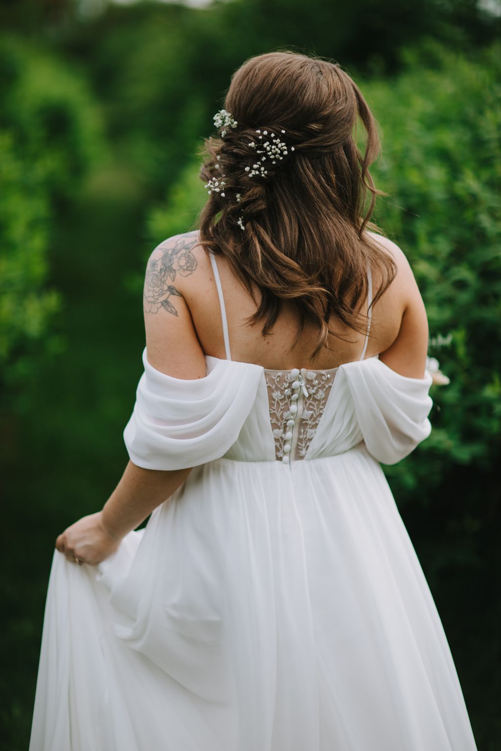 Bride in white off-shoulder dress with baby''s breath hair flowers, shown from behind against soft green garden background