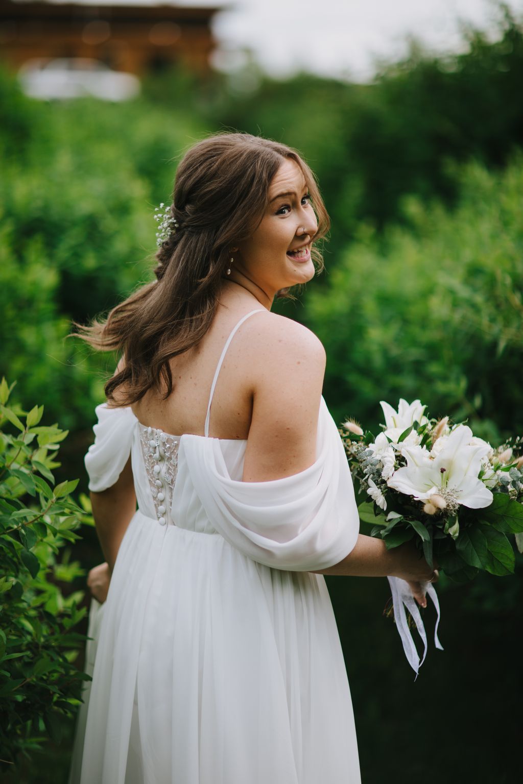 Bride in white flowing gown with baby''s breath in hair, smiling over shoulder in lush garden setting