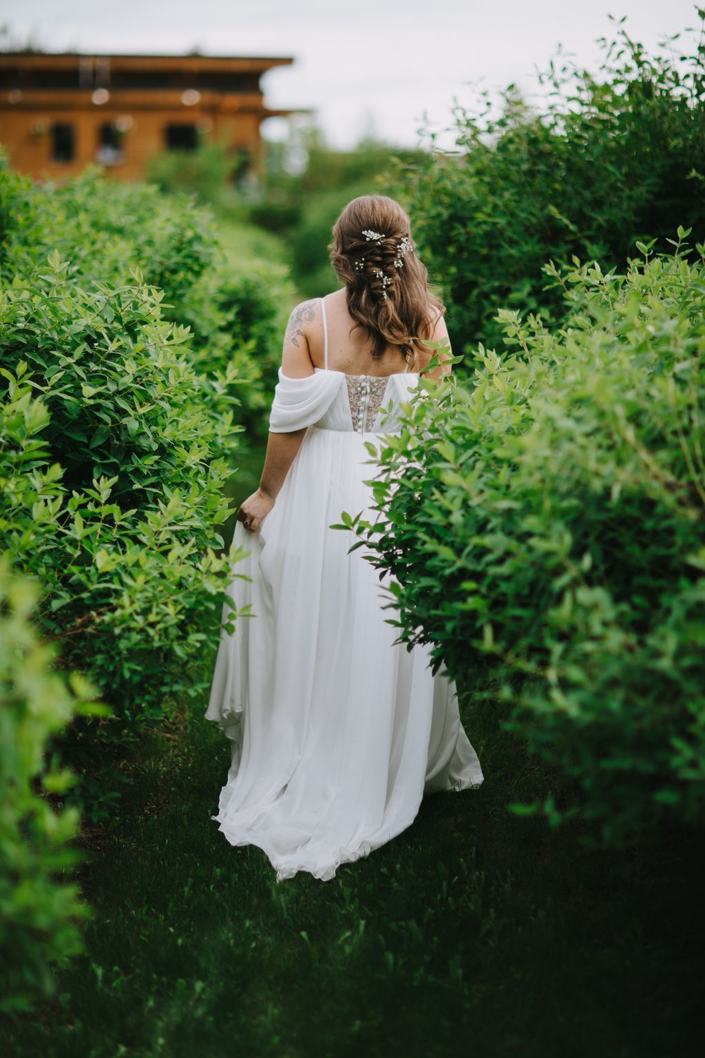 Bride in off-shoulder white gown with floral hair pins walks through lush green hedges toward a wooden structure