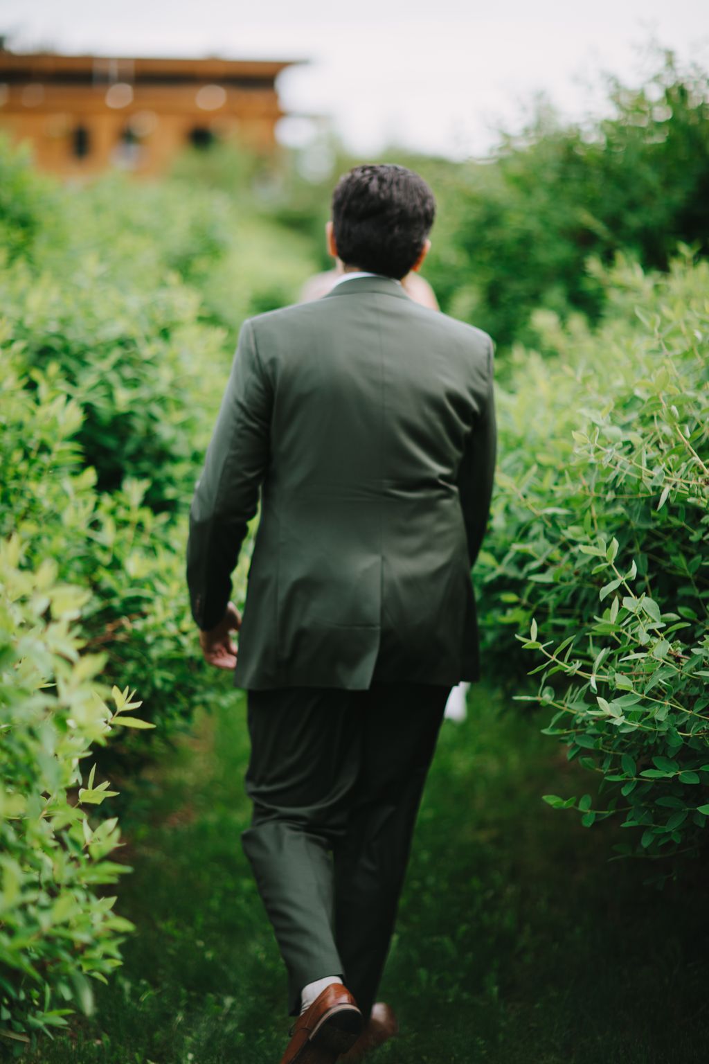 Groom in olive suit walking away through lush green garden with blurred brick structure in background