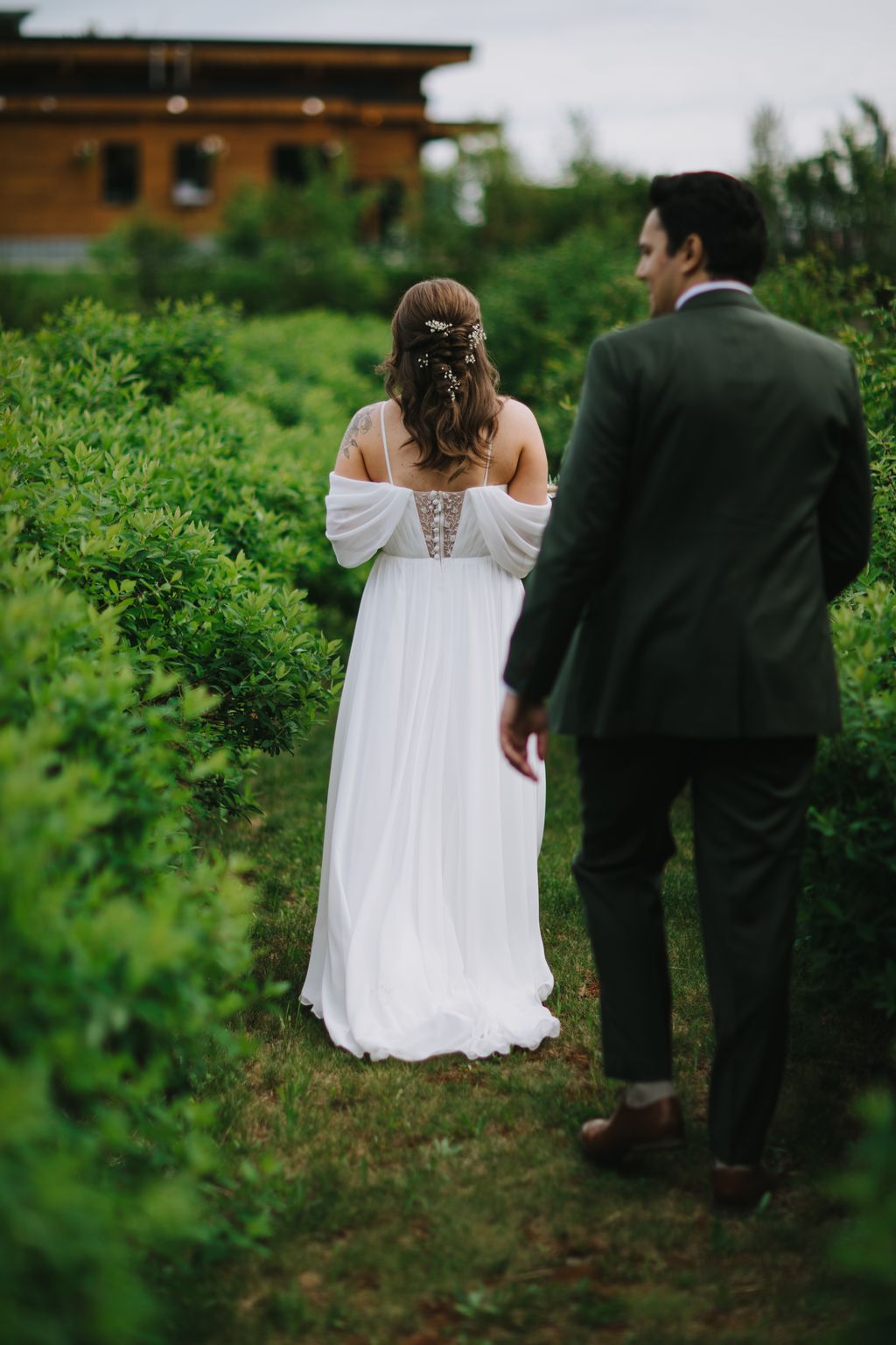 Bride in flowing white off-shoulder gown with beaded back stands in lush garden with groom in dark suit, soft natural light