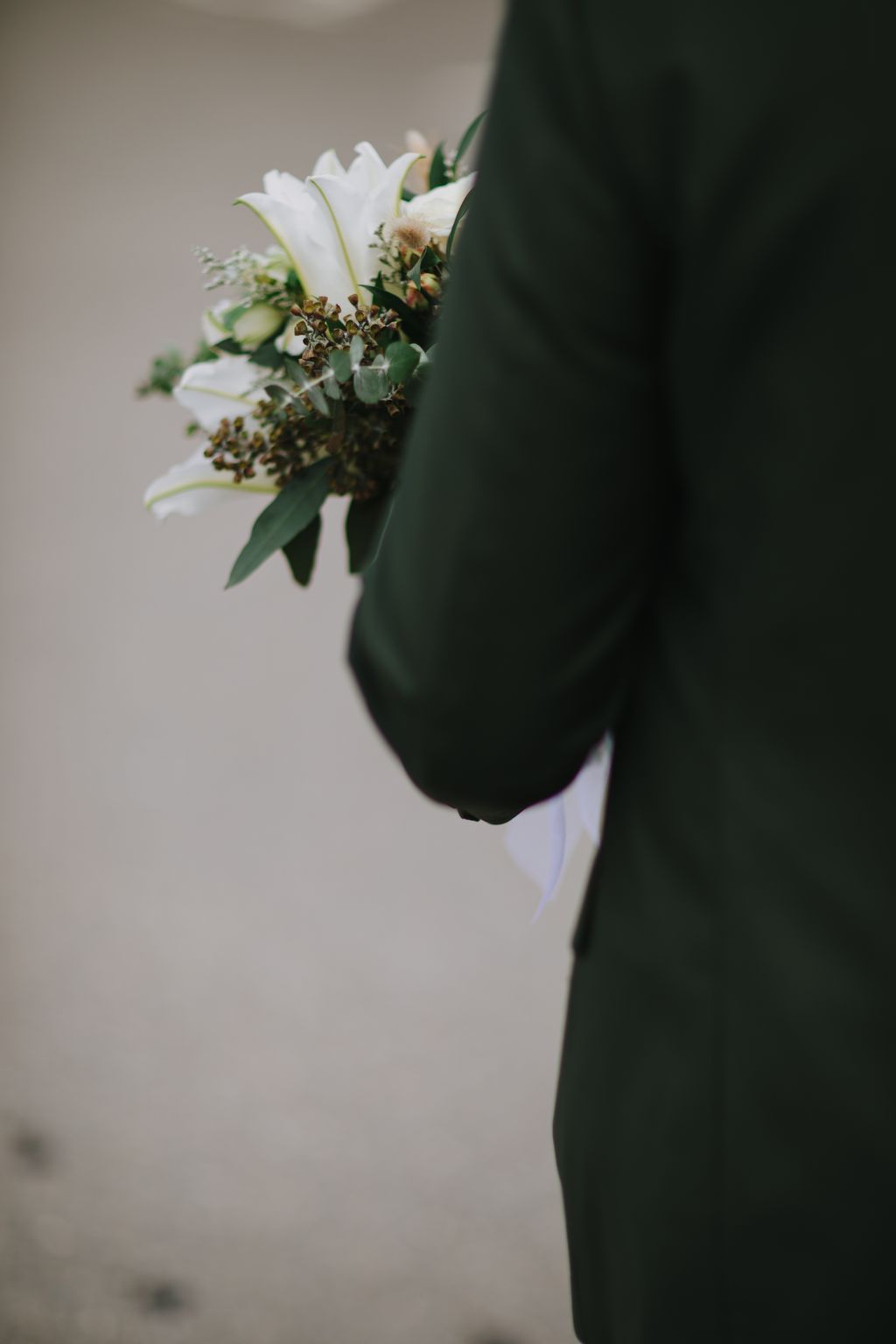 Groom in dark green suit holds white flower bridal bouquet with eucalyptus and dried botanicals against neutral background