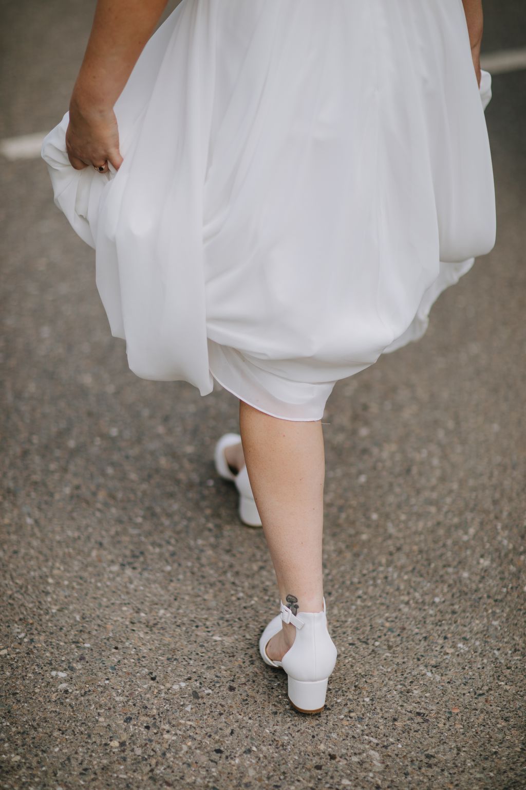 Bride in white flowing dress and heeled shoes walking on pavement, detail shot of lower body and legs