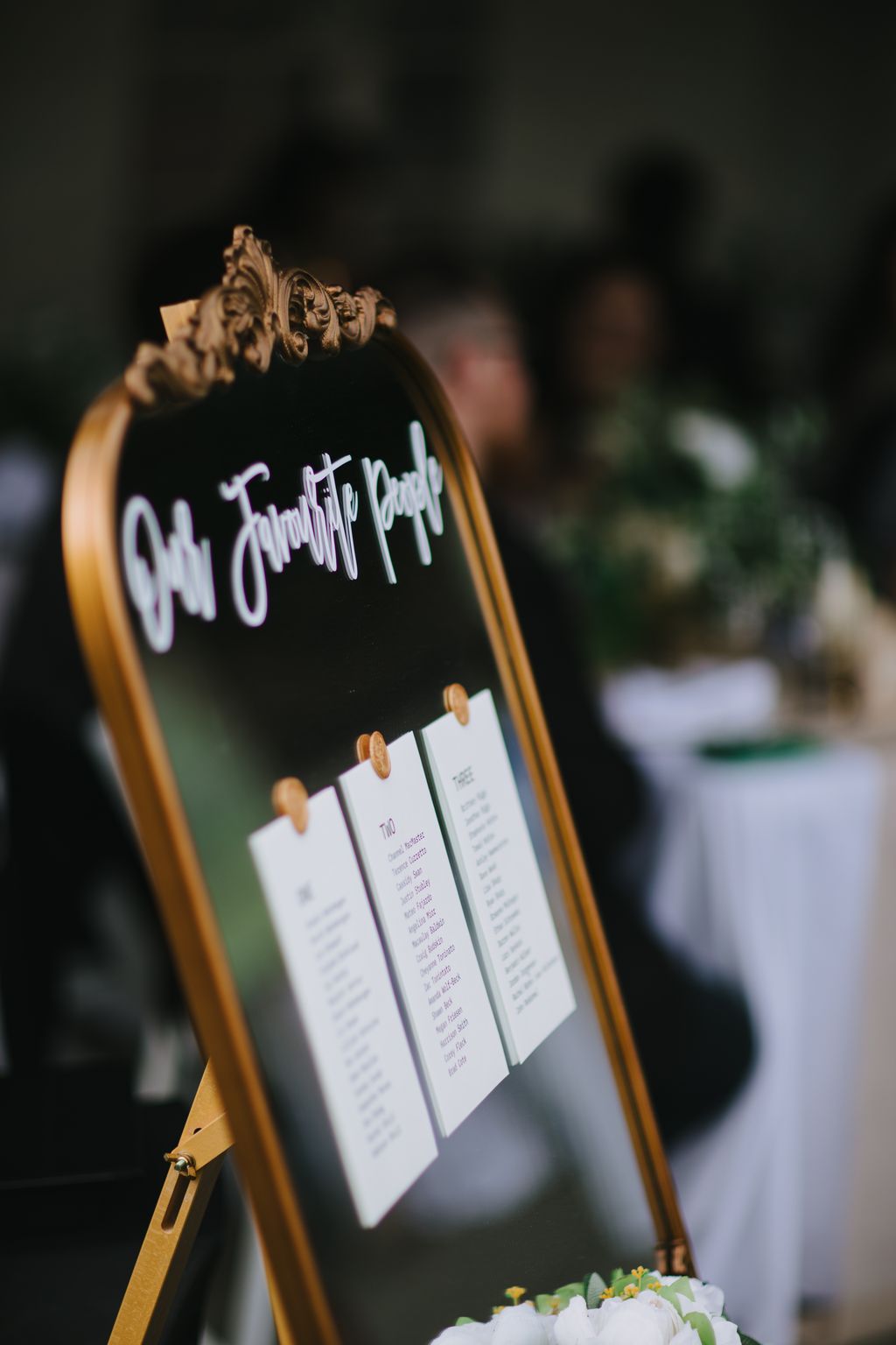 Ornate gold table card holder displaying white escort cards at an elegant wedding reception with soft blurred background