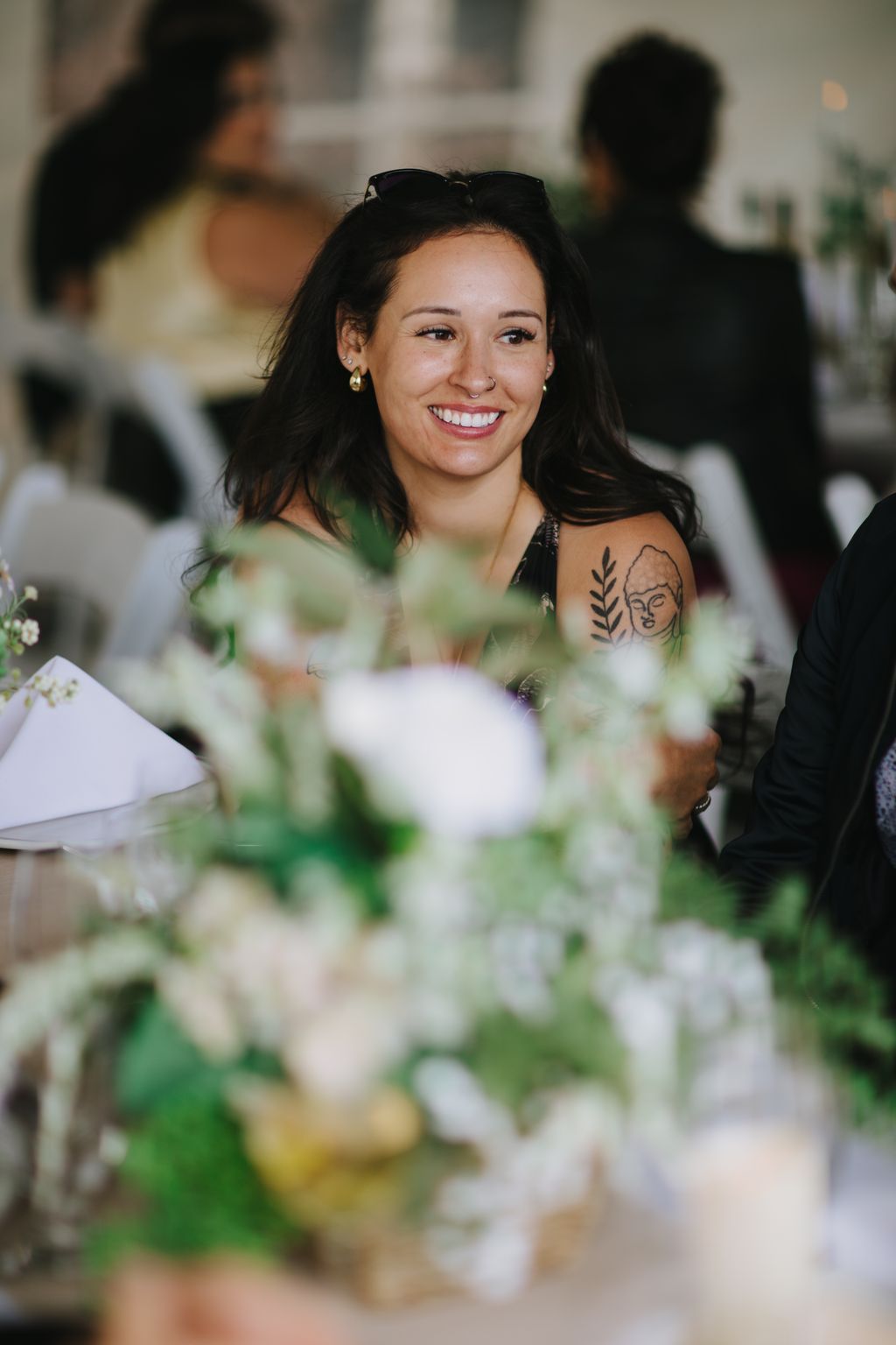 Woman with long dark hair smiling outdoors with white flowers in soft focus foreground and blurred background
