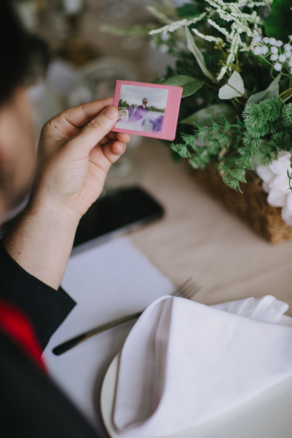 Hand holding pink instant photo of couple in lavender field, surrounded by greenery and white fabric