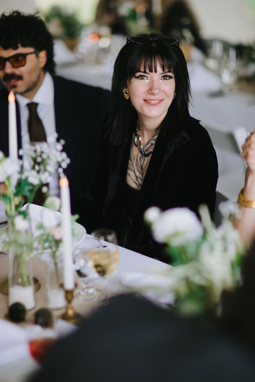 Woman with black hair and bangs smiling at elegant dinner table with wine glasses and soft bokeh background