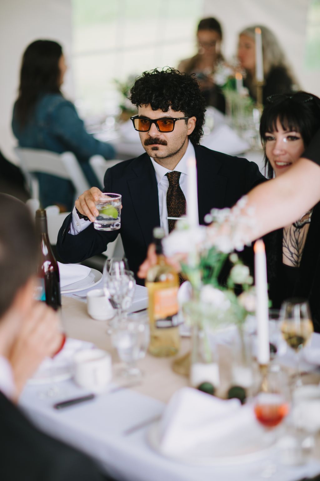 Groom in black suit and orange-tinted sunglasses holds a cocktail at an elegant table setting during reception