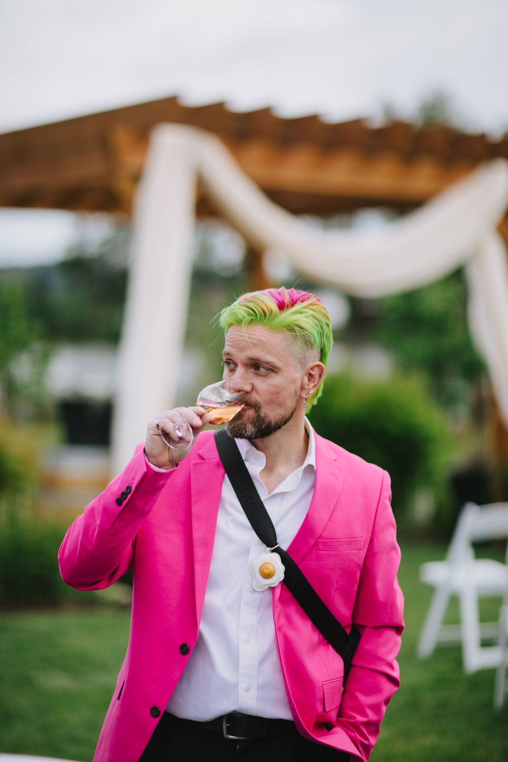 Groom in vibrant pink blazer sips champagne at outdoor wedding celebration with draped fabric backdrop