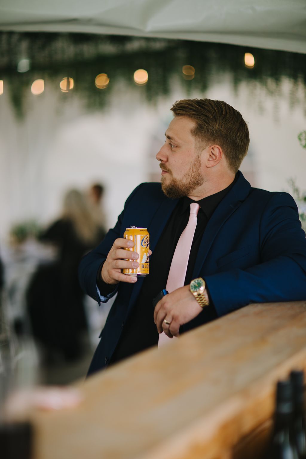 Man in navy suit and pink tie holding a beer can at an indoor event with warm bokeh lights in background