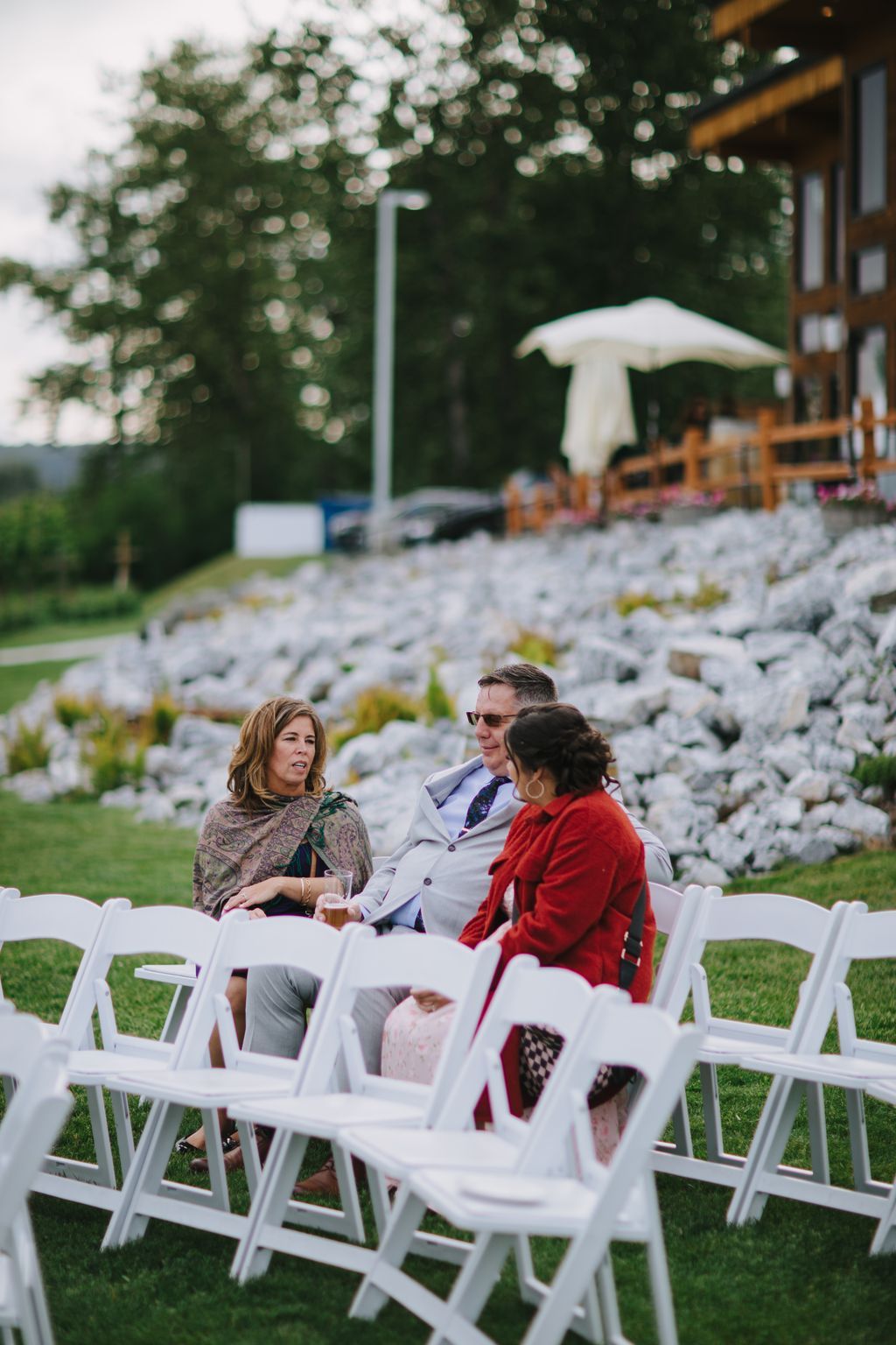 Three guests in elegant attire seated outdoors at dusk, conversing under string lights strung through trees, with white folding chairs and a landscape blurre…