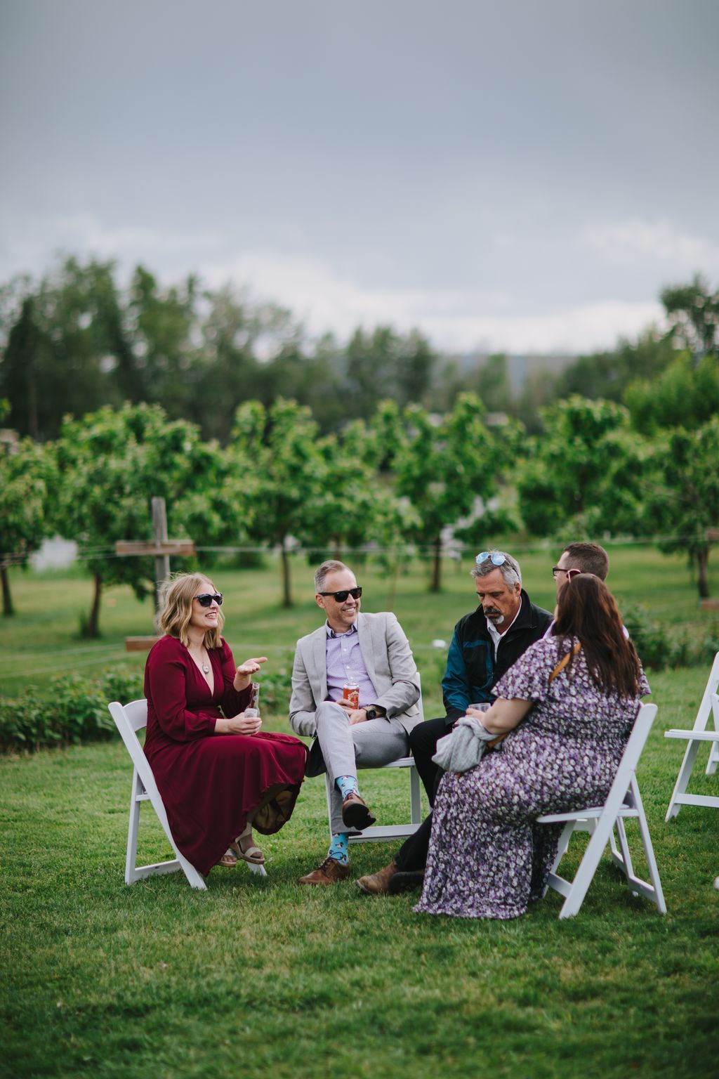 Guests in casual attire seated on chairs in a vineyard, conversing and smiling during an outdoor celebration with rolling hills and evergreens in the background