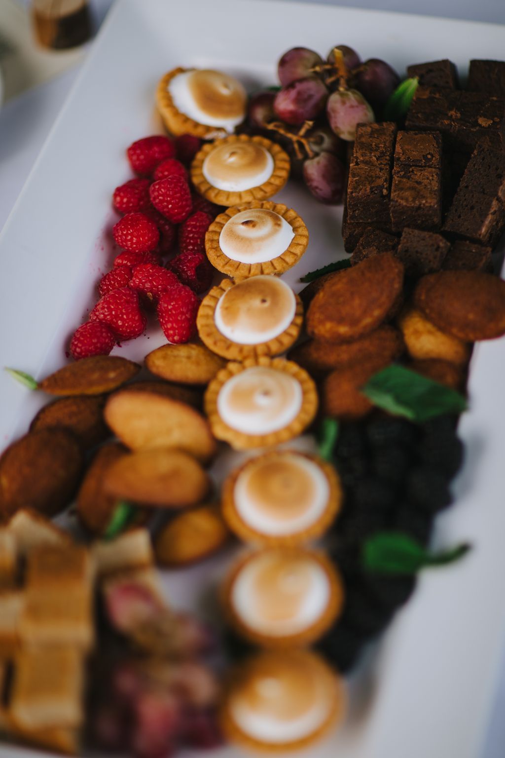 Elegant tartlets with torched meringue tops arranged with fresh raspberries and grapes on a white surface, showcasing refined dessert details