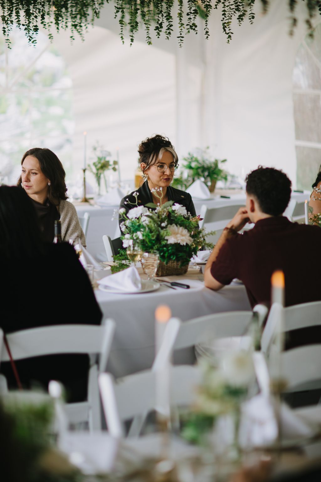 Woman in glasses and black jacket seated at white-draped table with white flowers and hanging greenery in bright, minimalist venue
