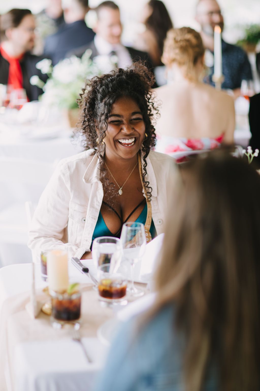 Woman with curly hair smiling at outdoor reception table with guests and drinks blurred in background