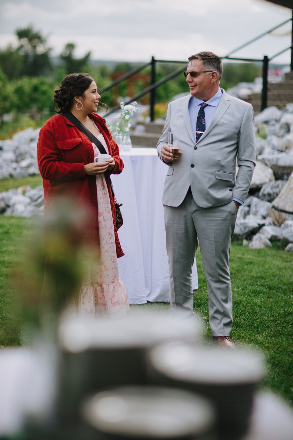Couple in elegant attire enjoying cocktails at an outdoor reception with scenic countryside backdrop and black gate details