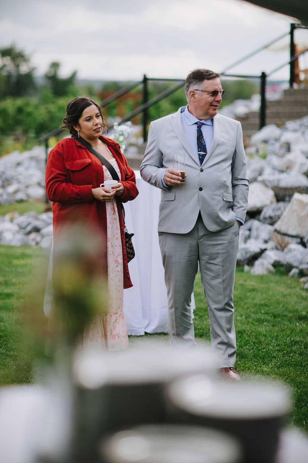 Couple in formal attire standing outdoors at rural wedding venue, woman in red wrap and floral dress, man in gray suit, overcast sky and green fields behind
