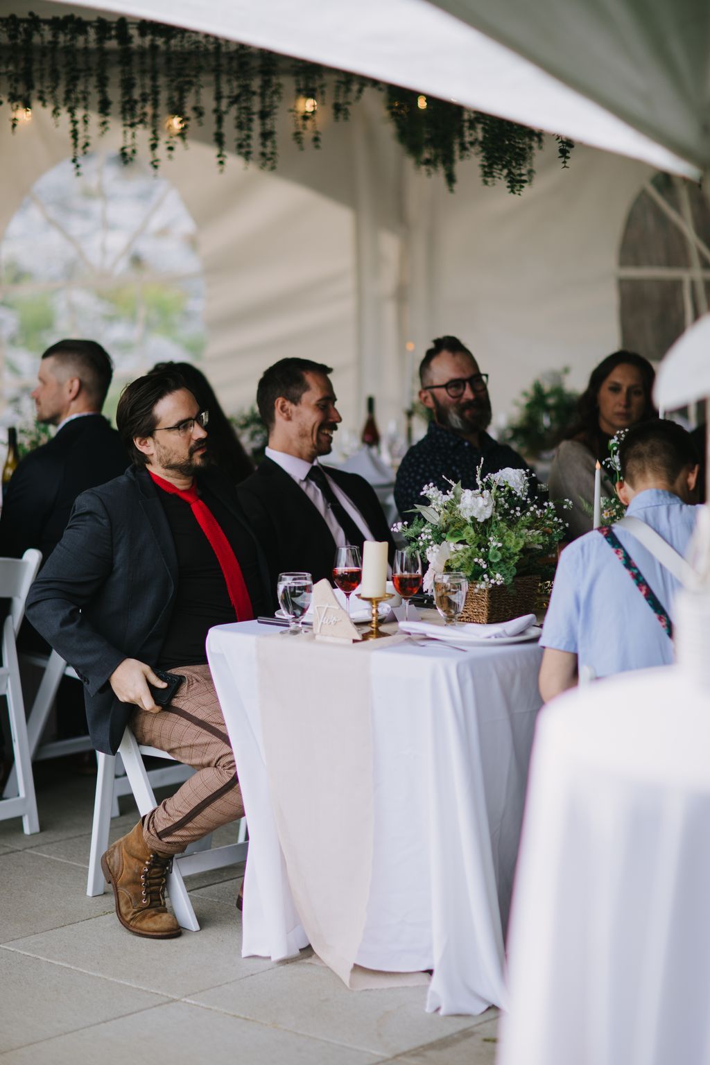 Guests seated at a white-draped table under a tent with hanging greenery and candlelight, smiling during an elegant reception