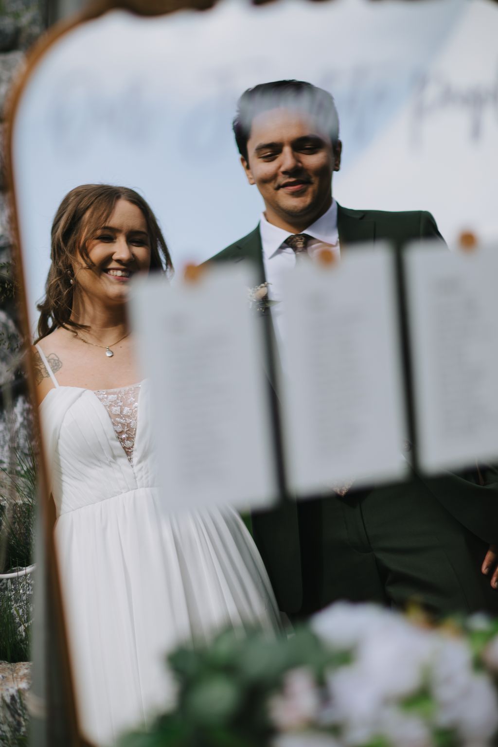 Groom in dark suit and tie smiles with eyes closed as bride in white dress stands beside him, shot through delicate veil with soft focus