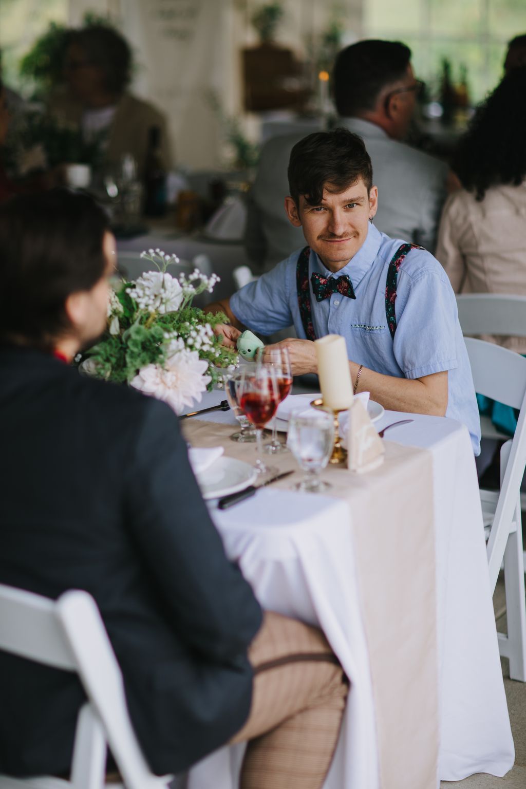 Groom in light blue shirt and patterned bow tie smiles while arranging white flowers at an elegant white-draped reception table
