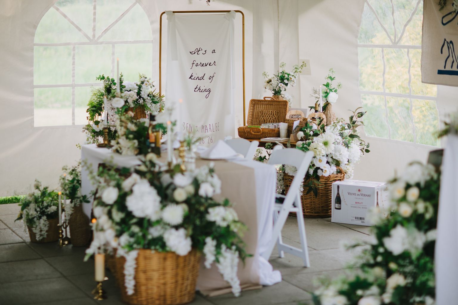 Romantic wedding ceremony backdrop featuring a white fabric banner reading "It''s a forever kind of thing" with couple''s names, surrounded by white flowers in…