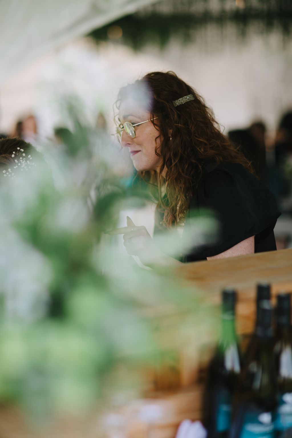 Woman with wavy brown hair and glasses wearing a sparkly hair clip, photographed in profile with soft green foliage in foreground and background